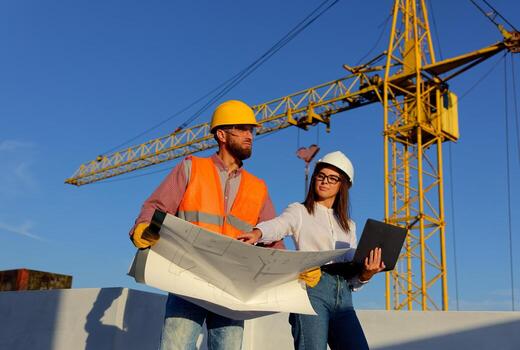 Construction workers discussing blueprint on building and laptop site with crane. photo
