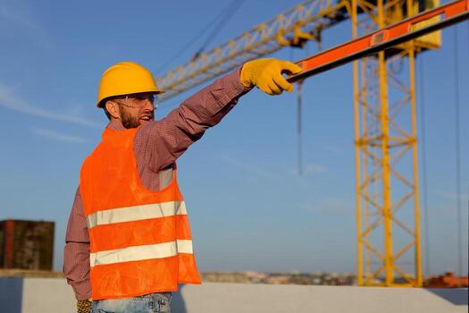 Construction worker in helmet holding level tool at building site with crane photo