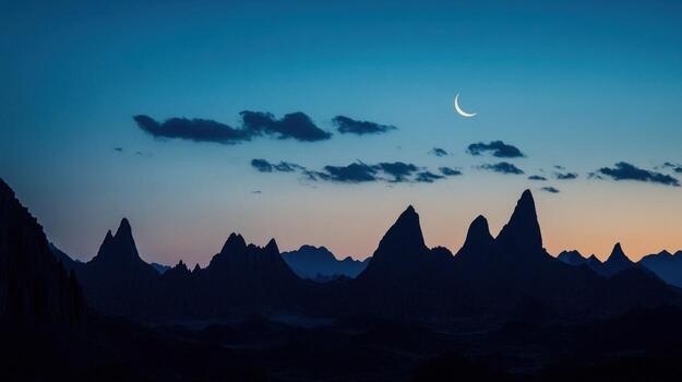 A crescent moon hangs in a twilight sky above a silhouetted mountain range, the peaks sharp against the deepening blue and purple hues of the evening. A few wispy clouds drift across the sky photo