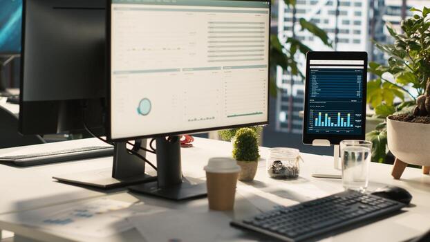 Modern workspace with analytical data dashboards displayed on devices in bright office environment. PC screen on desk with financial statistics graphs next to potted plants, jib down shot photo