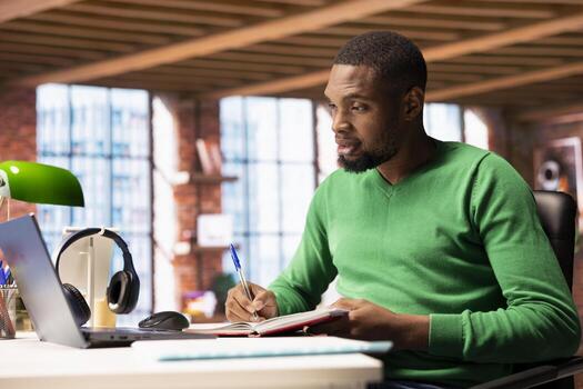 Person in home office analyzing data on notebook, writing info down on notepad, identifying trends. Freelancer using laptop compiling analytical reports, processing data, taking notes photo