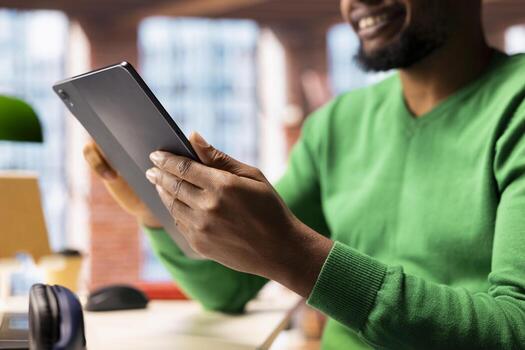 Smiling man at home taking break from work to look at social media feed on tablet, having fun. Happy remote worker in living room pausing tasks to use social network application on device, close up photo