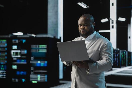 Software developer in data center using laptop, reading error logs, doing checkup to prolong hardware life span. Server hub computer scientist monitoring infrastructure rigs using notebook photo