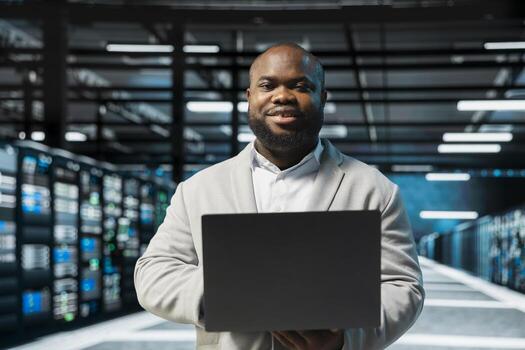 Portrait of upbeat server hub admin using laptop to fix network issues affecting storage arrays, doing backups. Jolly man in data center runs system diagnostic tests, monitoring thermal levels photo