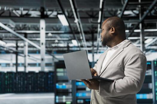 Programmer in data center using laptop, reading error logs, doing checkup to prolong hardware life span and avoid downtime. Server hub workplace employee monitoring infrastructure rigs using notebook photo