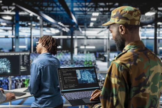 African american military staff uses AI tools for defense systems, working with IT developer in server room. Drone operator in military data center doing live drone monitoring on laptop. photo
