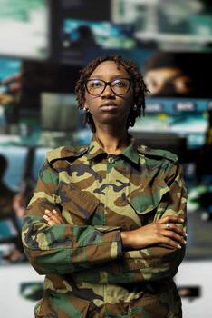 Portrait of officer in camouflage uniform standing with crossed arms against backdrop of digital surveillance screens in military command hub, showing discipline and readiness for defense operations photo