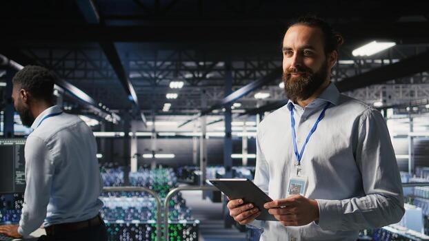 Cheerful data center admin doing maintenance tasks on tablet, ensuring optimal performance. Upbeat IT professional reviewing diagnostics and applying patches to server hub gear, photo