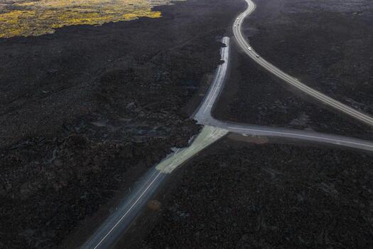 Aerial image of a road intersection in Iceland surrounded by dark lava fields, with a damaged section and hints of greenery in the upper left corner. photo
