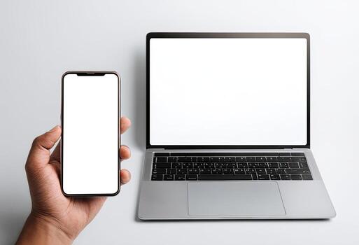 Hand holding phone next to open laptop with blank white screens on a white background photo