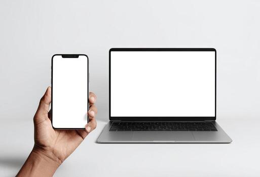 Hand holding a smartphone next to a laptop, both with blank white screens on a white surface photo