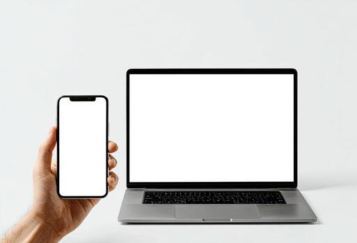 A hand holds a phone with a blank screen next to a laptop with a blank screen on white photo