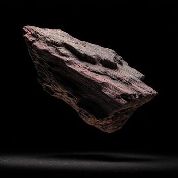 A solitary, substantial rock, dark gray with hints of rose, floats against a black backdrop. Its irregular shape suggests natural formation, not a manufactured object. A subtle shadow suggests a slight surface photo