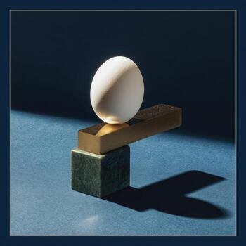 An egg balances atop a geometric stack, with a shadow cast against a blue backdrop photo