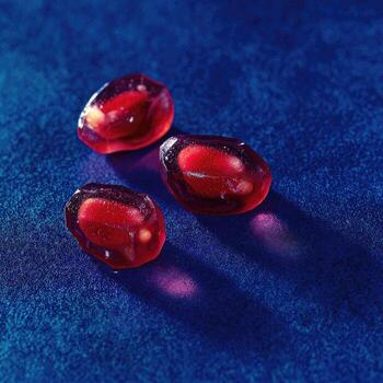 Three translucent ruby-red pomegranate seeds on a textured, deep blue surface, with light play photo