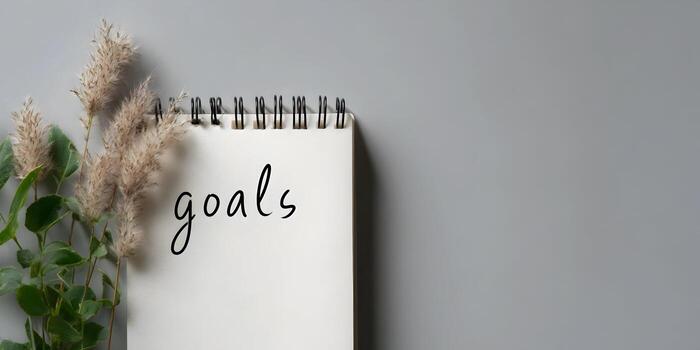 A simple top down view shows a blank page notebook with the word goals written in script alongside natural foliage on a solid grey surface photo