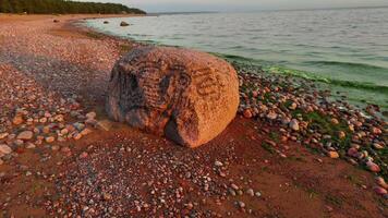 låg vinkel, närbild skott av en massiv rödbrun glacial- oregelbunden flyttblock med synlig gravyrer, vilar på en strand sammansatt av rödaktig sand och färgrik småsten, med grön vatten på grund av till alger video