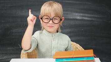 retour à école. content souriant élève dans rond des lunettes séance à le bureau. enfant a idée dans le salle de cours avec tableau noir sur Contexte. enfant garçon de primaire école. premier journée de automne. video