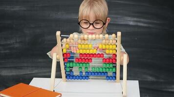 Back to school. Happy smiling pupil in round glasses sitting at the desk. Child playing colorful abacus in the classroom with blackboard on background. Kid boy from primary school. First day of fall. video