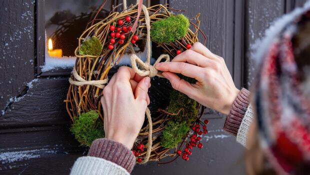 Hands tighten a jute knot on a natural winter wreath at a snowy door, with a candle glowing in the window photo