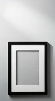 A simple black framed picture frame with a white matting rests against a textured light gray wall with subtle shadows photo