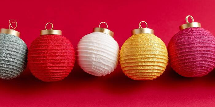 Five textured Christmas ornaments in gray, red, white, yellow, and pink are arranged in a row against a vibrant red background. Each ornament features a gold cap and hanging loop. photo