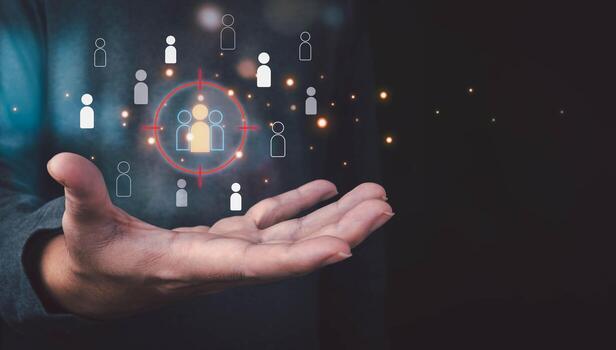 A person's hand holds a holographic interface, targeting a specific group of customer icons with a red crosshair, symbolizing marketing strategy and audience selection on a dark background. photo