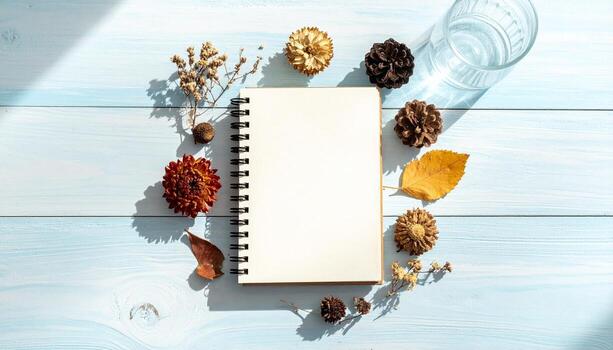 Flat lay of a blank notebook surrounded by autumn decor pine cones dried flowers and a glass of water on a light blue wooden table with soft shadow lighting photo