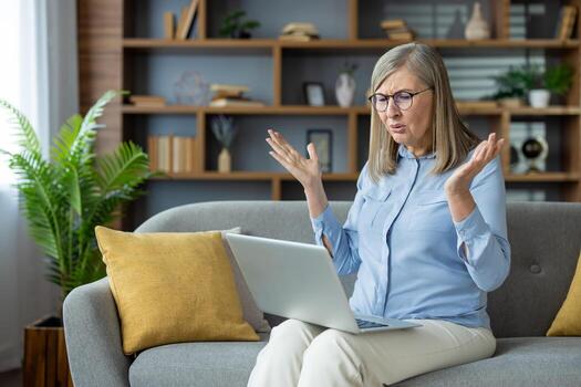 Senior woman displaying frustration during call on laptop while sitting on couch. Woman working home. Expression of confusion, irritation, and difficulty navigating computer program. photo