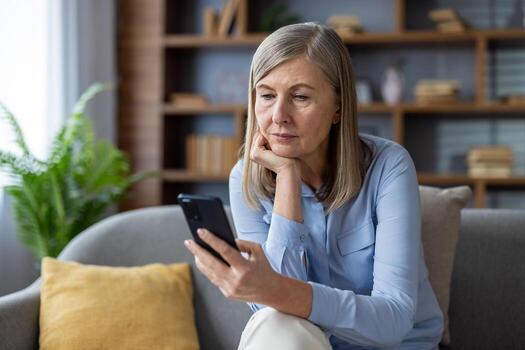 Mature woman using smartphone on sofa, showing thoughtful expression. Engaged in digital content interaction, casual living room setting, demonstrating technology use and depth of thought. photo