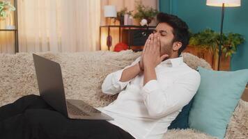 Indian young man at home, exhausted from work, falling asleep on the sofa with a laptop on his lap. Arabian guy tired lazy after long hours of work, resting on the couch, overwhelmed and drained video