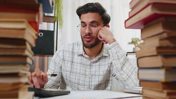 utmattad indisk affärsman granskning dokument rapporter medan använder sig av kalkylator arbetssätt hård i Hem kontor. betonade trött frilansare rörande huvud i förtvivlan omgiven förbi stackar av böcker på tabell. video