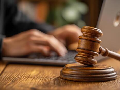 Close-up of wooden judge gavel on desk with blurred hands typing on laptop keyboard in background illustrating legal work and technology integration photo