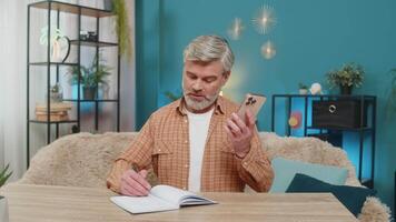 caucasico vecchio maturo uomo seduta a casa tavolo parlando su mobile Telefono altoparlante chiamata, scrittura Appunti nel taccuino. di mezza età tipo appare focalizzata, determinato, analizzando dati e In cerca di soluzioni video