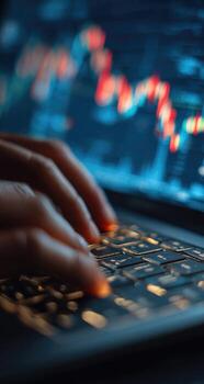 Close-up of hands typing on a laptop keyboard, blurred stock market graphs in the background photo