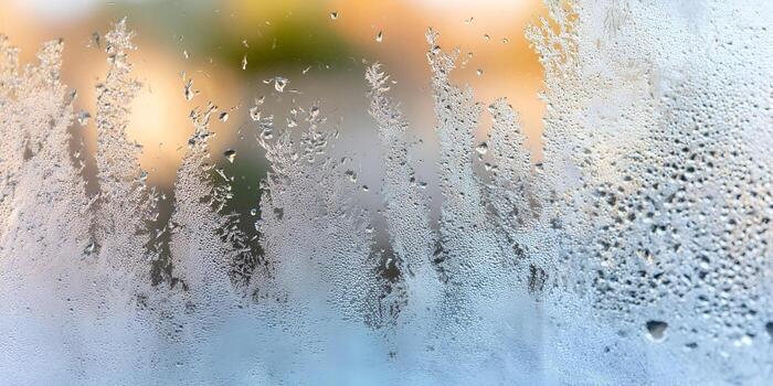 Intricate frost patterns and water droplets on a window glass creating an abstract winter texture with a blurred warm background photo