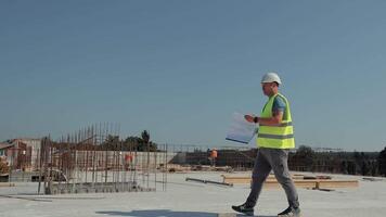 arkitekt granskning ritningar på modern konstruktion webbplats. ett arkitekt i en reflekterande väst och hjälm undersöker byggnad ritningar medan gående genom en video