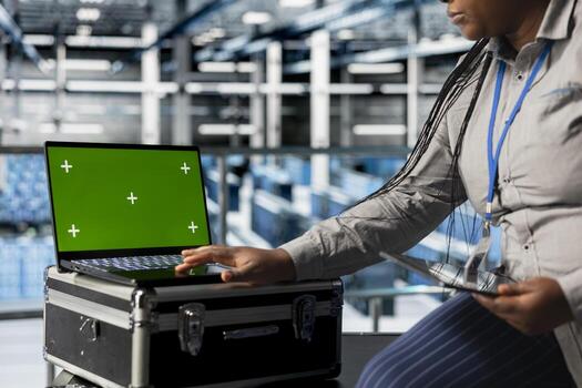 Mockup laptop next to female engineer in server hub implementing data backup and recovery solutions. African american data center employee using green screen notebook, optimizing network management photo