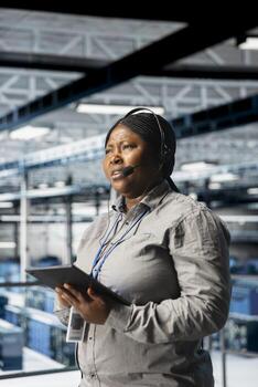 Data center engineer providing user technical support and solutions to users, using tablet to check diagnostics. African american woman in server hub wearing headset talking in mic with customers photo