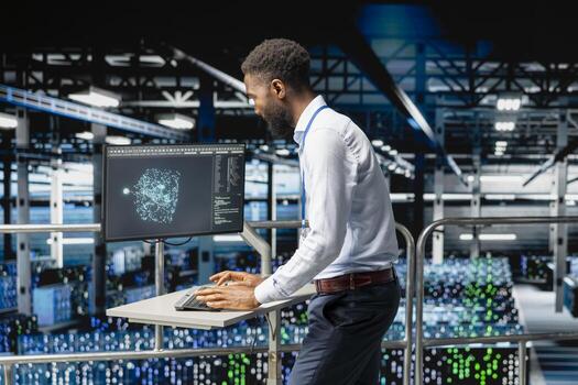 Black male specialist performing system maintenance on industrial platform in modern data center. IT expert using pc to do upgrades, resolving bugs and enhancing infrastructure stability. photo
