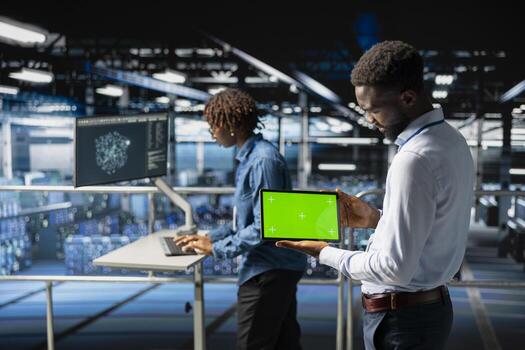 Black male technician using mockup screen on tablet to analyze neural network systems. System administrator uses green screen on tablet, reviewing LLM visualization on industrial platform. photo
