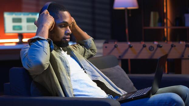 Self employed guy browsing the web and listening to music, having fun with songs on headset and completing tasks. African american man laying on sofa and relaxing in the living room. Camera B. photo