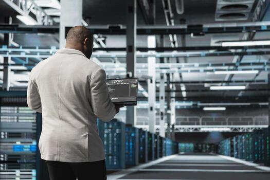 Programmer in data center typing on laptop, writing lines of code, compiling software patches to fix bugs. Server hub IT professional using programming scripts on laptop, conducting tests on equipment photo