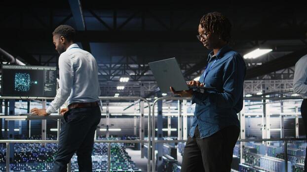 System administrators on data center industrial platform analyzing performance outputs on laptop. Server hub teamworking colleagues finetuning server rig configurations using notebook, photo