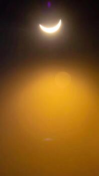 Celestial event captures the sun partially obscured by the moon, creating a crescent shape. Lens flare adds a vibrant golden glow and hazy atmosphere. An atmospheric halo is visible photo