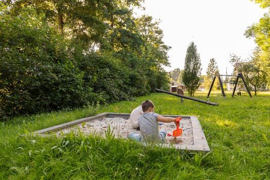 Two children are happily playing in a sandbox filled with sand, using small toys to dig and create, while surrounded by lush grass and trees in the warm afternoon light photo