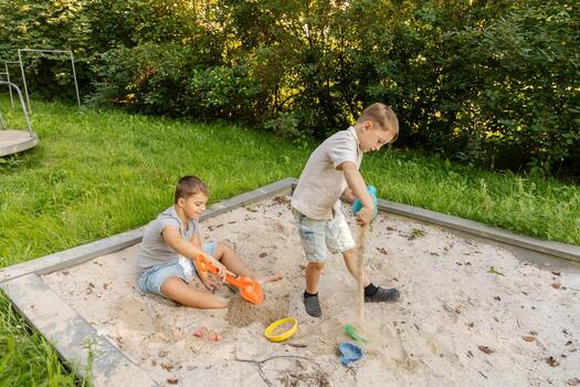 Two children are happily playing in a sandbox, digging with colorful shovels. Lush green grass and trees create a natural backdrop in a warm afternoon setting, enhancing their fun photo