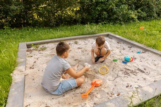 Two kids are playing in a sand pit surrounded by greenery. They are using colorful toys to build structures and enjoy each other's company on a sunny day photo