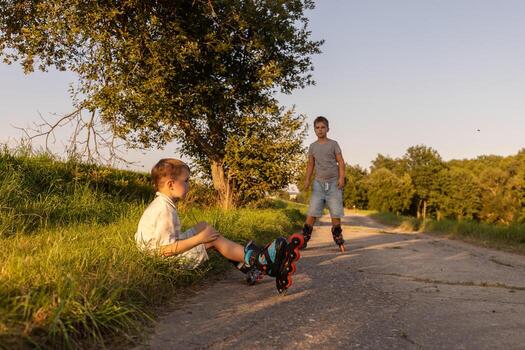 Two friends are inline skating on a warm afternoon. One child sits on the grass, while the other skates down the path, surrounded by trees and nature photo