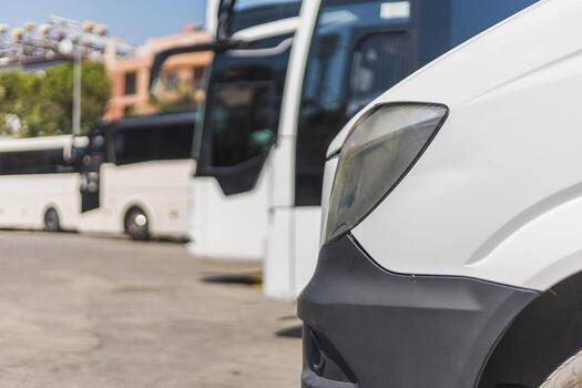 A close view of a white van shows its front while larger buses stand parked in the background. The scene is set at a busy transport station on a sunny day photo
